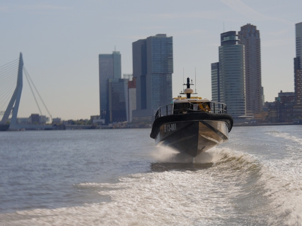 Image of the Artemis EF-12 Workboat foiling in the Port of Rotterdam with the skyline and bridge in the background. The vessel is in the colour of the brand.