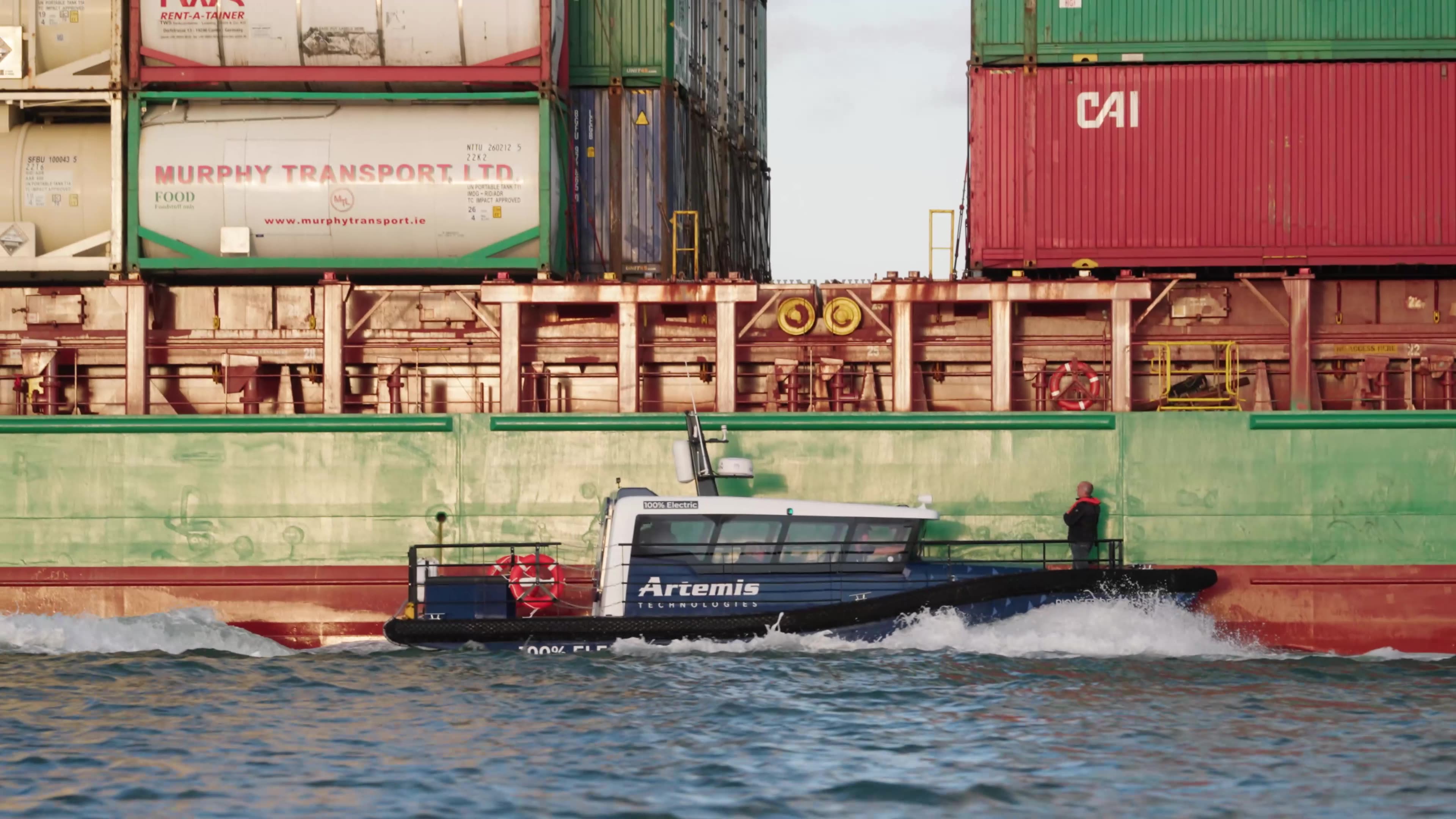 Artemis eFoiler® 12 metre vessel making contact with a large red container ship with a person at the bow of the smaller vessel near to boarding position