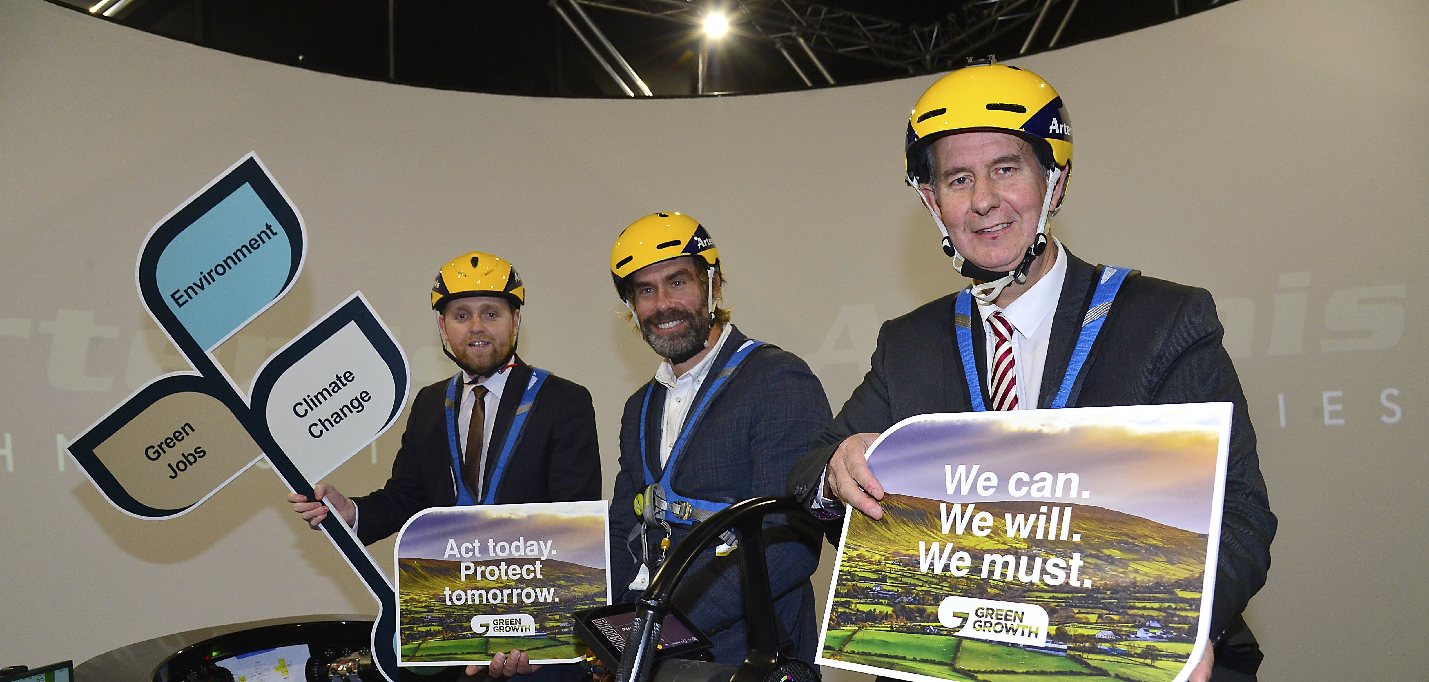 Iain Percy with two MPs on the Artemis Technologies Simulator holding signs about climate change and green growth