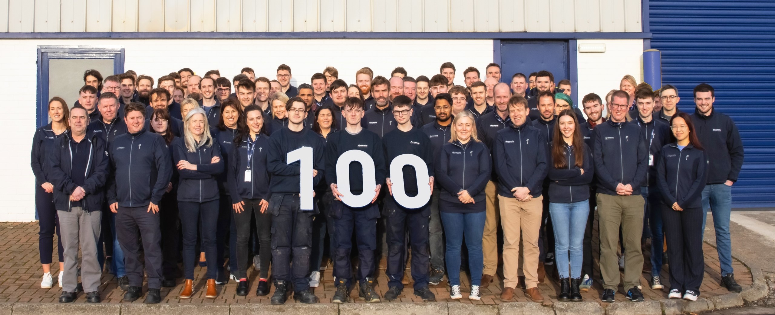 Group shot of 100 employees holding 100 sign outside of Artemis Technologies' Manufacturing facility