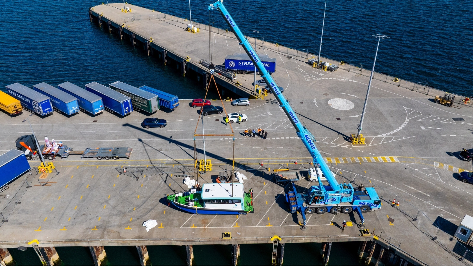 Vessel on pier in Orkney with a blue crane. The vessel has a white body a blue hull and a green deck. It is a sunny day.