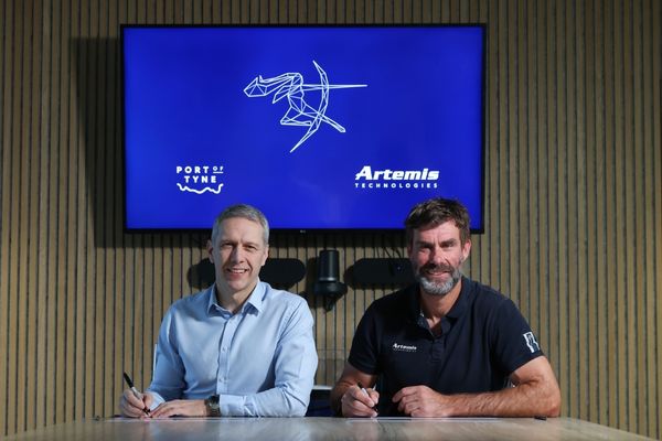 Two men sitting at a table signing while smiling and a TV in the background with the Port of Tyne and Artemis Technologies logo in the background