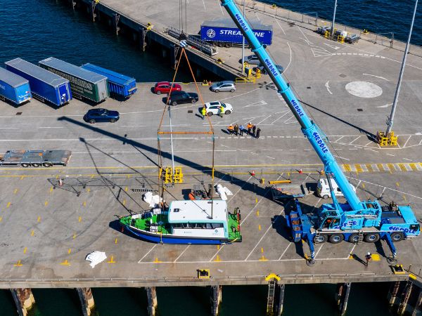 Image shows the Artemis EF-12 Workboat XL on the pier at Orkney from above with a blue crane lifting it and people in hard hats and hi-vis vests. It is a sunny day and the vessel is blue, white and green.