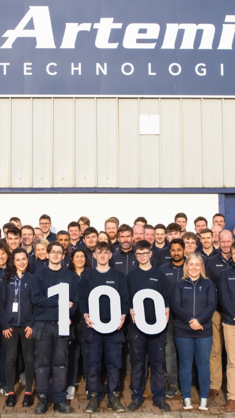 group of 100 artemis employees holding 100 sign outside manufacturing facility 